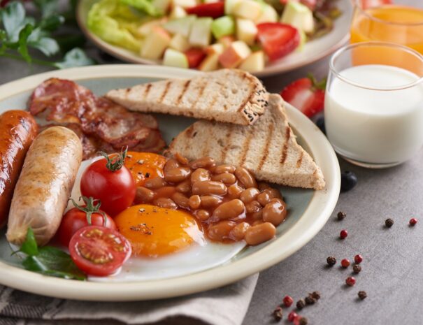 Traditional full English breakfast with fried eggs, sausage, tomato, beans, toast and bacon on a plate, Fresh fruit and vegetable salad, Milk and orange juice on stone background.